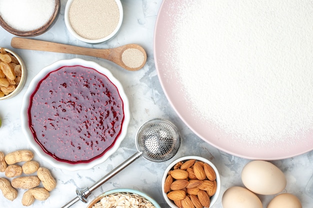 Top view powdered flour on plate bowls with flour oats almonds jam walnut eggs wooden spoon on table