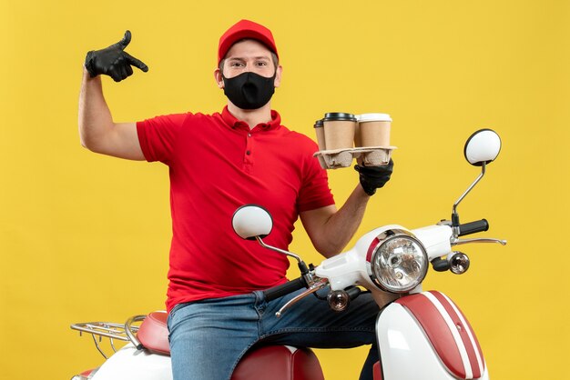 Top view of proud delivery guy wearing uniform and hat gloves in medical mask sitting on scooter showing orders