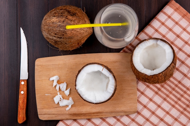 Top view of pulps of coconut on a wooden kitchen board with knife and a glass of water on checked tablecloth and black surface