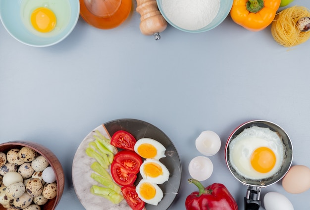 Top view of quail eggs on a wooden bowl with boiled halved eggs on a plate with slices of tomato with fried eggs on a frying pan on white background with copy space