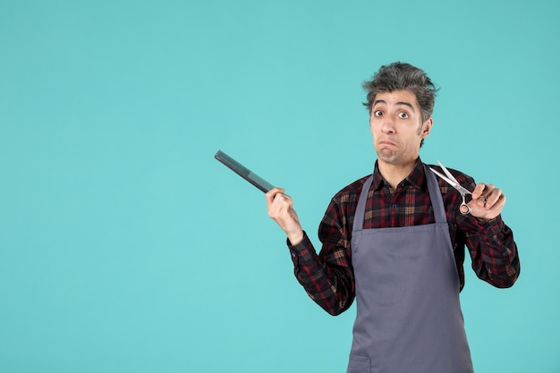 Top view of questioning male barber wearing gray apron and holding comb scissor on blue color background