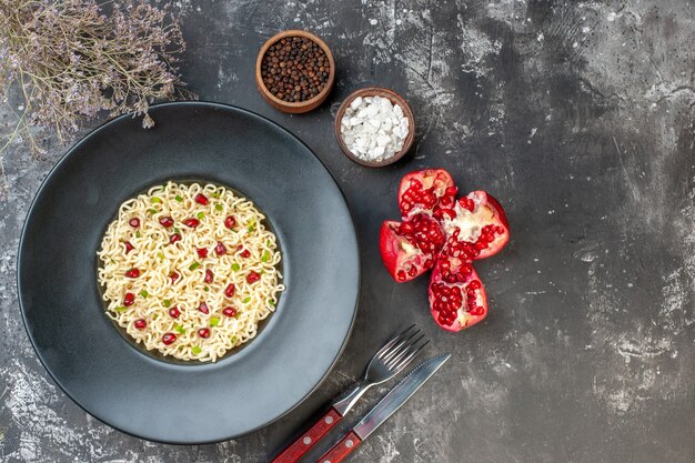 Top view ramen noodles on round plate a fork and knife black pepper in bowl pomegranate on dark table free space