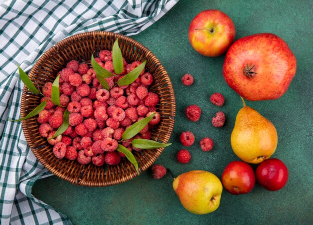 Top view of raspberries in basket on plaid cloth and pattern of pomegranate peach apple plum on green surface