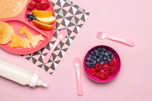 Top view of raspberries and blueberries in bowl with baby bottle and food