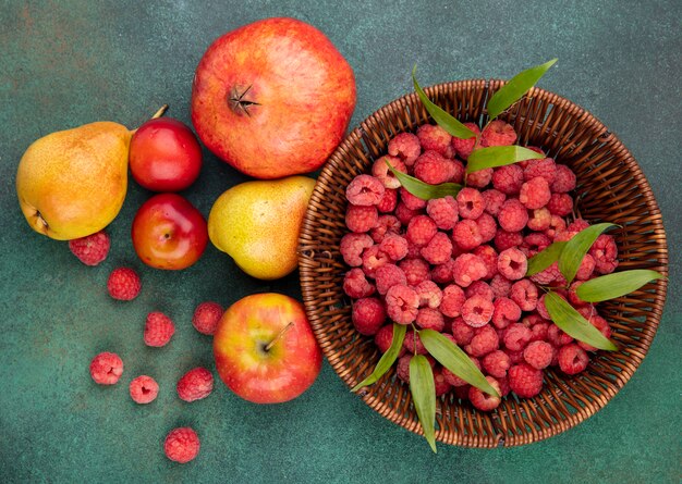 Top view of raspberries in bowl and pattern of pomegranate peach apple plum on green surface