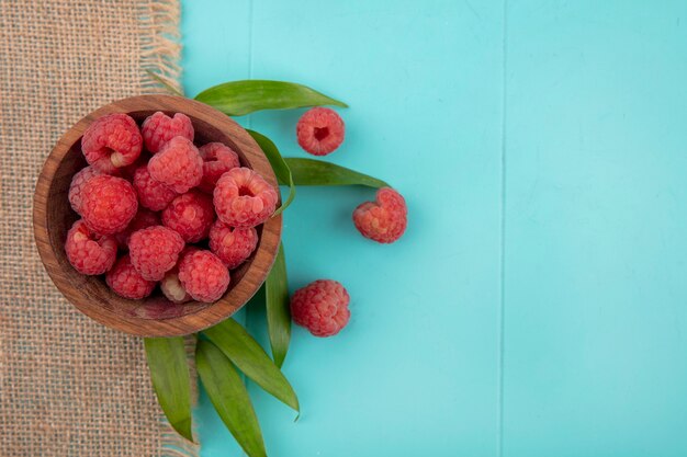 Top view of raspberries in bowl on sackcloth with leaves on blue surface