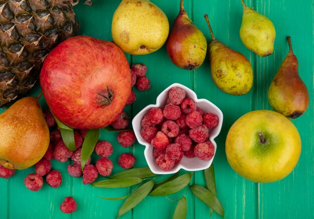 Top view of raspberries in bowl with pomegranate pineapple peach and apple with leaves around on green surface