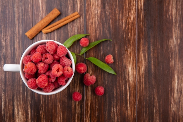 Top view of raspberries in cup with cinnamon and leaves around on wooden surface