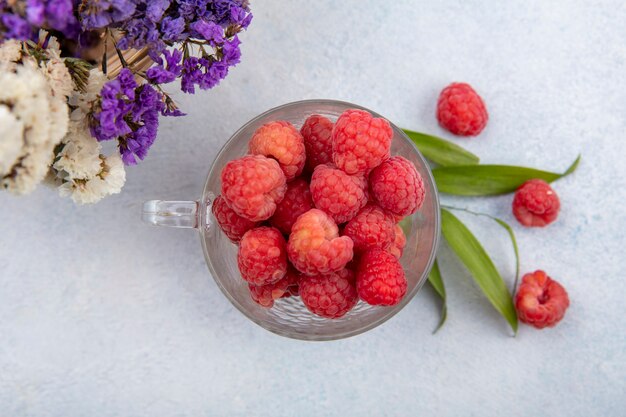 Top view of raspberries in glass cup with flowers and leaves on white surface