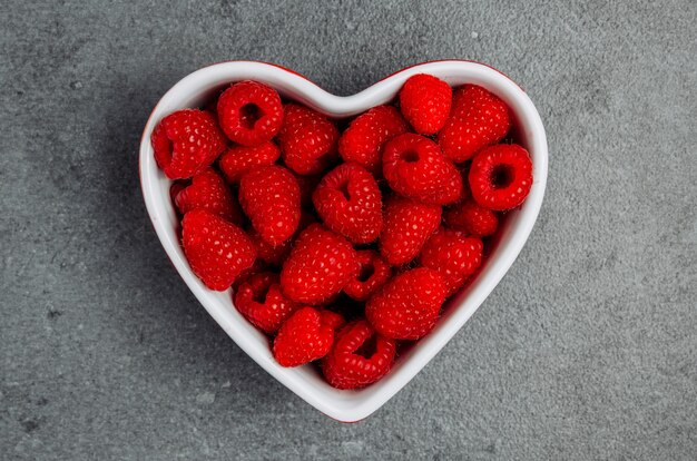 Top view raspberries in heart shaped bowl on gray textured background.