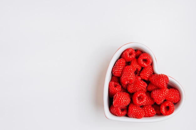 Top view raspberries in heart shaped bowl on white background. horizontal space for text