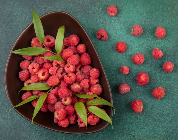 Top view of raspberries and leaves in bowl and pattern of raspberries on green surface