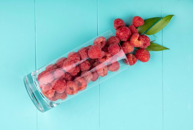 Top view of raspberries spilling out of glass with leaves on blue surface