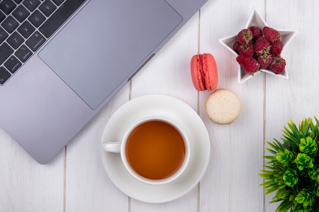 Top view of raspberries with a cup of tea macaroons and a laptop on a white surface