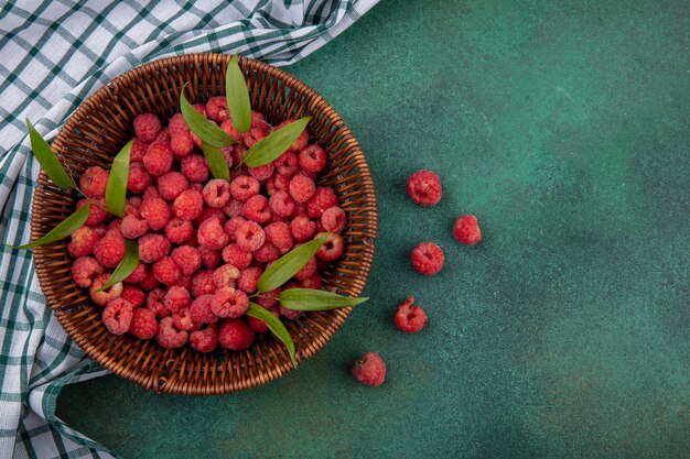 Top view of raspberries with leaves in basket on plaid cloth and green surface