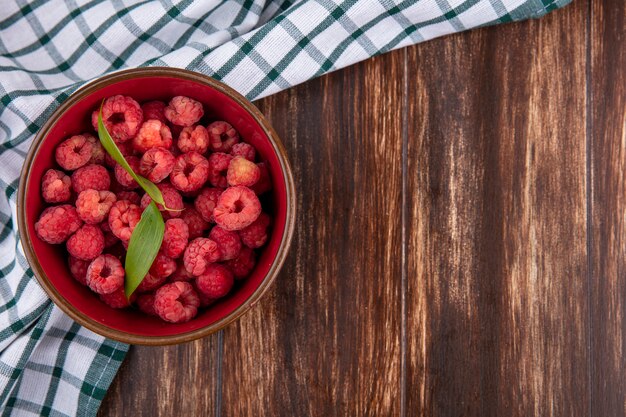 Top view of raspberries with leaves in bowl on plaid cloth on wooden surface