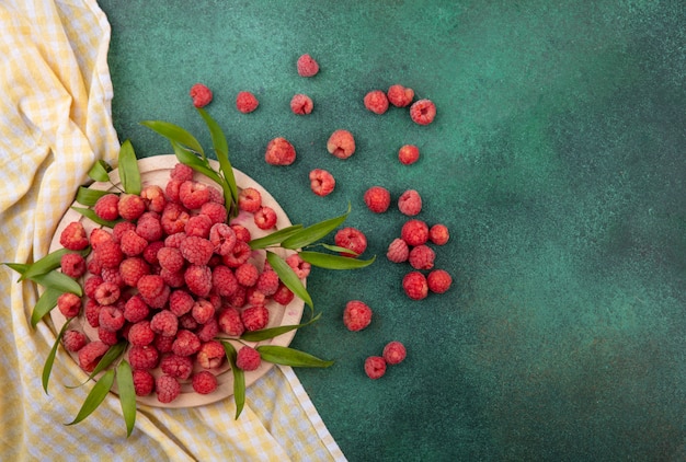 Top view of raspberries with leaves on cutting board on plaid cloth and on green surface