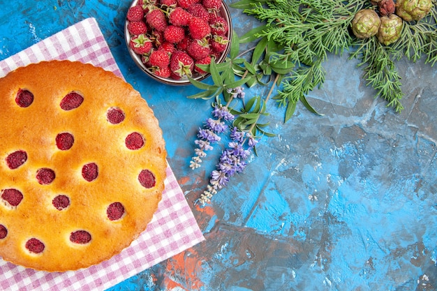 Top view of raspberry cake on kitchen towel bowl with raspberries pine tree branch on blue surface