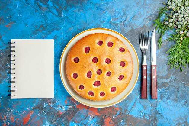 Top view of raspberry cake on oval plate cones fork knife a notebook on blue surface