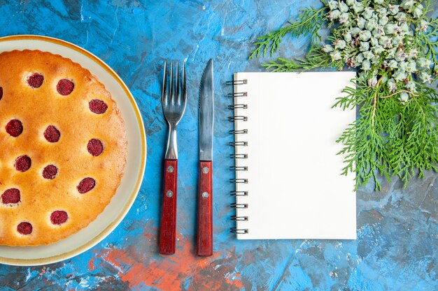 Top view of raspberry cake on oval plate cones fork knife a notebook on blue surface