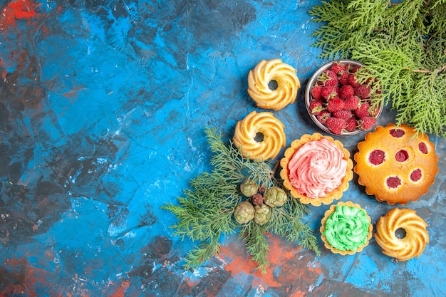 Top view of raspberry cake, small tarts, biscuits, bowl with berries and tree branches on blue surface