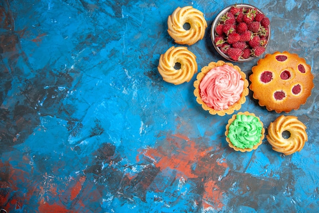 Free photo top view of raspberry cake, small tarts, biscuits and bowl with raspberries on blue pink surface