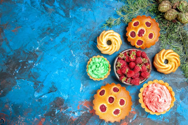 파란색 표면에 딸기와 나무 딸기 케이크, 비스킷, 작은 타르트, 콘 및 그릇의 상위 뷰