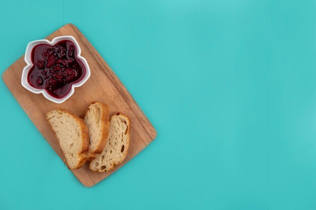 Top view of raspberry jam in bowl and sliced baguette on cutting board on blue background with copy space