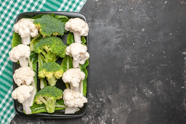 Top view raw broccoli and cauliflower on black rectangular plate on green and white checkered napkin on dark surface with copy space