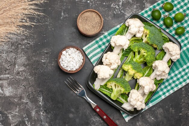 Top view raw broccoli and cauliflower on black rectangular plate on green and white checkered napkin fork sea salt pepper feykhoas on dark surface free space
