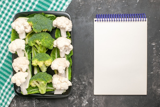 Top view raw broccoli and cauliflower on black rectangular plate on green and white checkered napkin a notebook on dark surface