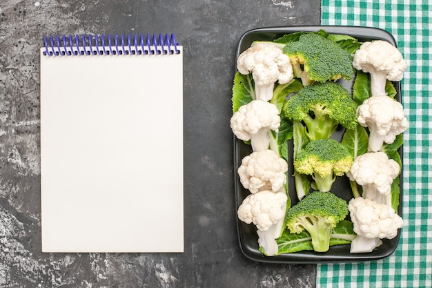 Foto gratuita vista dall'alto broccoli crudi e cavolfiore sulla zolla rettangolare nera sul tovagliolo a quadretti verde e bianco un taccuino sulla superficie scura