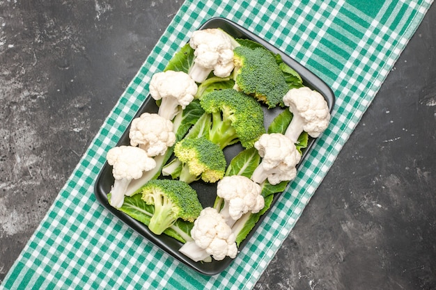 Top view raw broccoli and cauliflower on black rectangular plate on green and white checkered tablecloth on dark surface