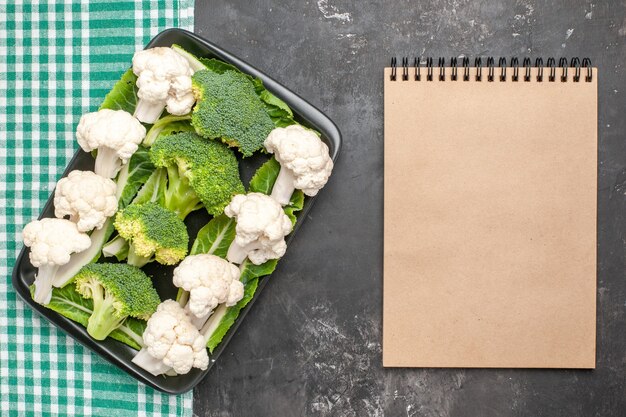 Top view raw broccoli and cauliflower on black rectangular plate on green and white checkered tablecloth a notebook on dark surface