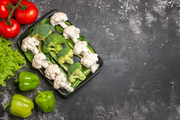 Top view raw broccoli and cauliflower on black rectangular plate tomatoes green bell peppers lettuce on dark surface free place