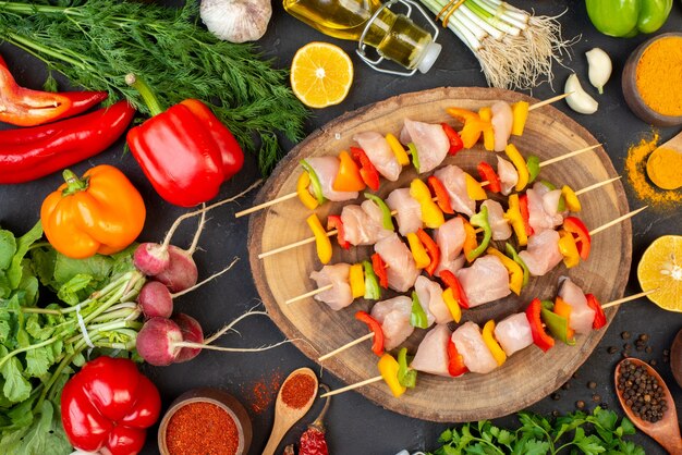 Top view of raw chicken skewers on natural wood board vegetables oil bottle spices on dark table