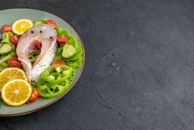 Top view of raw fish and fresh chopped vegetables lemon slices spices on a gray plate and cutlery set on the right side on black surface