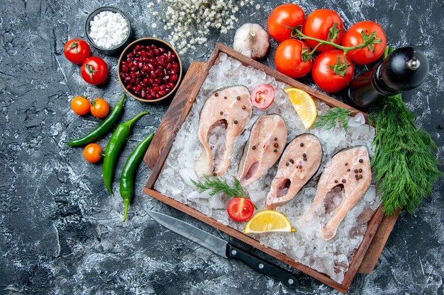 Вид сверху кусочки сырой рыбы со льдом на деревянной доске, зеленые острые перцы, миски с семенами пемаграната, морская соль, помидоры, укроп на столе