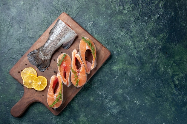 Top view of raw fishes lemon slices greens pepper on wooden cutting board on dark table