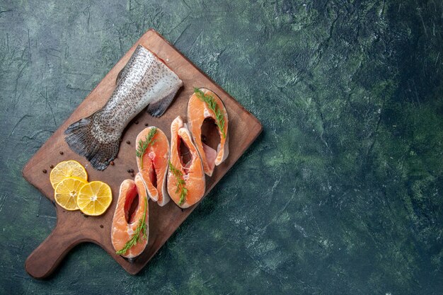Top view of raw fishes lemon slices greens pepper on wooden cutting board on dark table