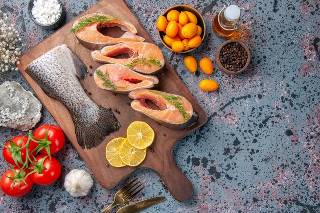 Top view of raw fishes lemon slices greens pepper on wooden cutting board and foods flower on blue black colors table