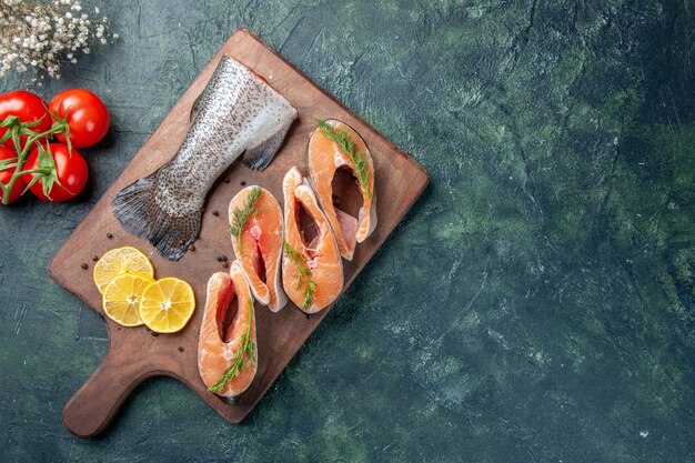 Top view of raw fishes lemon slices greens pepper on wooden cutting board tomatoes on dark table