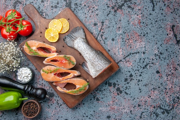 Top view of raw fishes lemon slices greens pepper on wooden cutting board and vegetables on blue black colors table