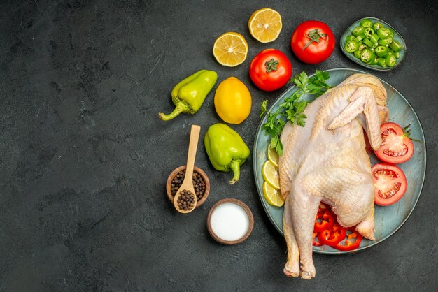 Top view raw fresh chicken inside plate with greens lemon and vegetables on dark background