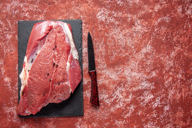 Top view of raw fresh red meat on black board and knife on the right side on oil pastel red background with free space