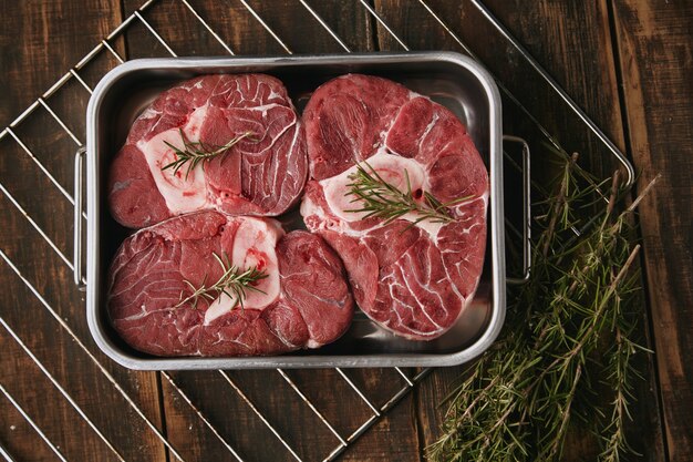 Top view of raw meat in steel pan ready to cook in oven. Spices around: pepper, garlic, rosemary, onions. salt. Sause pot. warm tones.