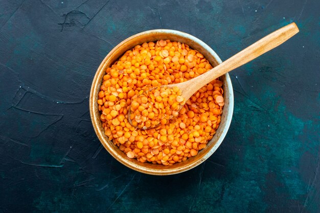 Top view of raw orange lentil inside round pot with wooden spoon on dark blue surface