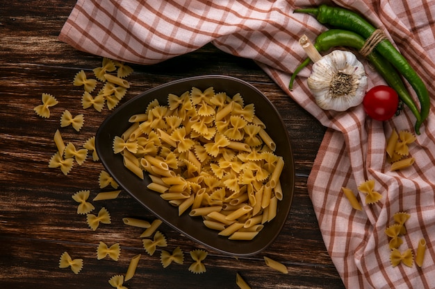 Top view of raw pasta in a bowl with garlic tomato and chili pepper on a checkered towel on a wooden surface