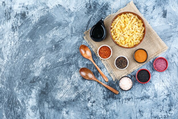 Top view raw pasta in bowl with spices, scoop, wooden spoons on plaster and piece of sack background. horizontal