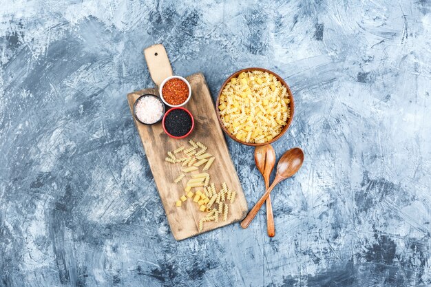 Top view raw pasta in bowl with wooden spoons, spices on grey plaster and cutting board background. horizontal